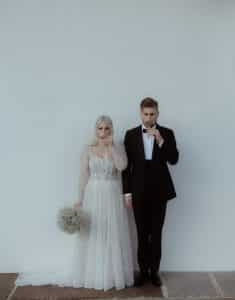 Ein Paar steht vor einer weißen Wand und feiert seine Hochzeit mit Gläsern in der Hand, während es in die Kamera blickt. Die Braut strahlt in ihrem weißen Kleid und hält einen Strauß kleiner Blumen in der Hand, und der Bräutigam ergänzt sie in einem klassischen schwarzen Smoking mit Fliege.