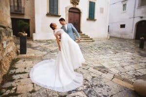 Ein lächelndes Paar im Hochzeitskleid tanzt in einem rustikalen, gepflasterten Innenhof und verkörpert die Freude der Hochzeitsplanung. Der Bräutigam trägt einen hellblauen Anzug, während die Braut ein weißes Kleid mit langer Schleppe trägt, wunderschön eingerahmt von dem weißen Gebäude und seiner großen Holztür.