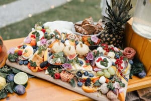 Eine farbenfrohe Wurstplatte, perfekt für eine Hochzeit, mit einer Auswahl an Käse, Früchten, Nüssen und Fleisch. Dekoriert mit frischen Blumen, Trauben, geschnittenen Feigen, Beeren und einer ganzen Ananas. Cracker und Brot stehen daneben auf einem Holztisch im Freien.