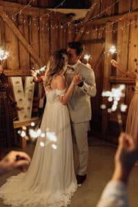 Ein Paar tanzt während seiner Hochzeit in einer warm beleuchteten Scheune, umgeben von Menschen mit Wunderkerzen. Die Braut trägt ein weißes Kleid und der Bräutigam einen hellen Anzug. Funkelnde Lichterketten hängen darüber und verstärken die romantische Atmosphäre dieser wunderschön geplanten Feier.