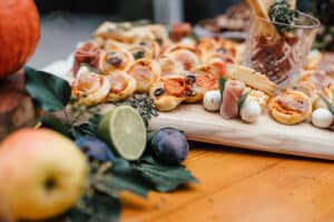 Ein Holzbrett mit verschiedenen Häppchen, perfekt für eine Hochzeitsfeier, darunter Minipizzas, Oliven und Prosciutto-Rollen. Frische Früchte wie Äpfel, Pflaumen und eine Limette sind mit Blattgemüse als Garnitur verteilt. Ideal, um Ihr Hochzeitsplanungserlebnis zu bereichern.