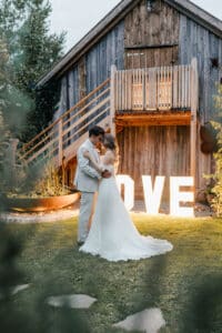 Braut und Bräutigam tanzen romantisch auf einer Wiese vor einem rustikalen Holzgebäude und feiern ihre Hochzeit. Umgeben von Grün umarmen sie sich im Schein großer Leuchtbuchstaben, die „LOVE“ buchstabieren.