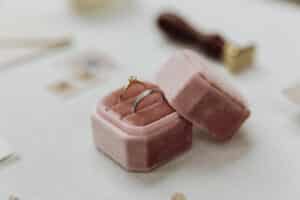 Ein einfacher goldener Verlobungsring mit einem kleinen Diamanten liegt in einer offenen Schmuckschatulle aus rosa Samt auf einer weißen Oberfläche, perfekt für diejenigen, die gerade mitten in der Hochzeitsplanung stecken. Im Hintergrund sind verschiedene verschwommene Gegenstände zu sehen.