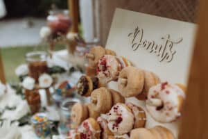 Auf einem Holzbrett mit der Aufschrift „Donuts“ sind verschiedene Donuts ausgestellt, einige mit Zuckerguss und Streuseln, andere ohne Zuckerguss. Im Hintergrund sorgen Gläser mit Bonbons und weißen Blumen für eine festliche Hochzeitsstimmung, perfekt für jede Hochzeitsfeier.