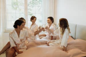 Fünf Frauen in Satinroben sitzen auf einem Bett und stoßen an. Umgeben von sanftem Licht und einem Fenster im Hintergrund feiern sie in einer warmen und fröhlichen Atmosphäre, perfekt für Festlichkeiten vor der Hochzeit.