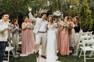 Ein fröhliches Brautpaar feiert seine Hochzeit und schreitet durch den Gang im Freien. Der Bräutigam winkt fröhlich, während die Braut einen Blumenstrauß hält. Brautjungfern in rosa Kleidern und Gäste applaudieren, umgeben von üppigem Grün und weißen Stühlen.