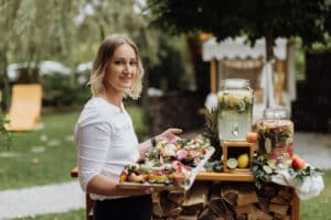 Eine Person mit schulterlangem Haar steht im Freien und verkörpert die Essenz der Hochzeitsplanung. Sie hält ein Holztablett voller bunter Speisen. Dahinter stehen auf einem Holzständer in einer üppigen Gartenlandschaft Gläser mit Getränken und schaffen eine idyllische Kulisse, die perfekt für jede Hochzeitsfeier ist.