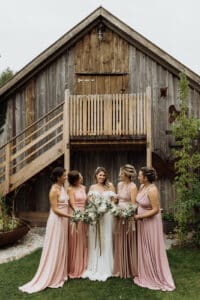 Eine Braut in einem weißen Kleid steht mit vier Brautjungfern in passenden rosa Kleidern da und hält Sträuße aus Grünpflanzen und weißen Blumen in den Händen. Sie posieren elegant vor einer rustikalen Holzscheune, die sich perfekt für jede romantische Hochzeitskulisse eignet, da eine Treppe zu einer Dachbodentür führt.