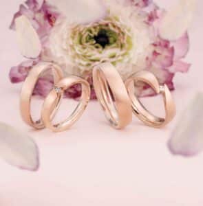 Vier elegante Roségoldringe, perfekt für eine Hochzeit, werden vor einem zartrosa Hintergrund präsentiert, eingerahmt von zarten Blütenblättern und einer blühenden Blume in der Mitte. Jeder Ring hat ein elegantes, modernes Design, einige zeigen kleine Diamantakzente, ideal für diejenigen, die heiraten möchten.