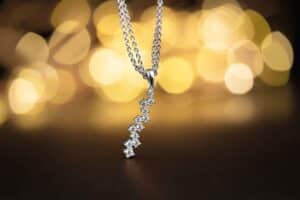 Eine silberne Halskette mit einer vertikalen Reihe kleiner, funkelnder Diamanten schwebt vor einem verschwommenen, sanft leuchtenden Hintergrund aus warmem, goldenem Licht und verleiht jeder Hochzeitsfeier Eleganz.