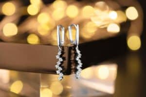 Elegante Silberohrringe mit kaskadierenden diamantähnlichen Steinen werden auf einer reflektierenden Oberfläche präsentiert und verleihen Ihrer Hochzeit einen Hauch von Glamour. Die warmen, verschwommenen Bokeh-Lichter im Hintergrund schaffen eine luxuriöse und festliche Atmosphäre für ein unvergessliches Hochzeitserlebnis.