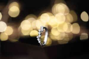 Ein silberner Ring mit funkelnden Diamanten ruht auf einer dunklen Oberfläche, vor einem verschwommenen Hintergrund aus goldenen Lichtern, und schafft eine warme und elegante Atmosphäre, die perfekt ist, um die Freude einer Hochzeit zu feiern.