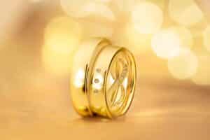 Eine Nahaufnahme von zwei goldenen Eheringen auf einem weichen, goldenen Hintergrund fängt die Essenz einer Hochzeit ein. Ein Ring ist mit drei kleinen Diamanten verziert, während der andere ein Unendlichkeitssymbol mit winzigen Diamanten trägt. Das Bild ist sanft beleuchtet und schafft eine warme, elegante Atmosphäre, die perfekt für die Hochzeitsplanung ist.