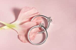 Zwei Silberringe mit kleinen Diamanten liegen auf einem zartrosa Hintergrund neben einem rosa Blütenblatt und fangen die Essenz von Eleganz und Romantik ein. Perfekt für Hochzeitsfeiern, ihr minimalistisches Design spricht moderne Paare an, die in Hochzeitsglück versunken sind.