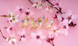 Eine Sammlung goldener Ringe mit bunten Edelsteinen wird elegant zwischen rosa Kirschblüten auf einem pastellrosa Hintergrund präsentiert und verleiht Ihrer Hochzeitsplanung einen romantischen Ton.