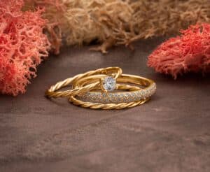 Ein Set eleganter Goldringe, perfekt für eine Hochzeit, sitzt anmutig auf einer strukturierten Oberfläche. Ein Ring ist mit einem schillernden Diamanten verziert, während ein anderer mit kleinen Diamanten geschmückt ist, alles umgeben von rosa korallenartigen Verzierungen, ideal für alle Hochzeitsplanungsbedürfnisse.