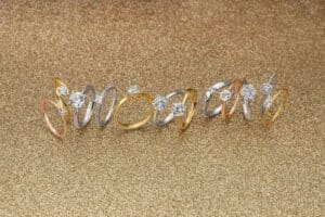 Eine Reihe eleganter Ringe mit funkelnden Diamanten ziert eine schimmernde Goldoberfläche, perfekt für alle, die eine Hochzeit planen. Diese Ringe sind in verschiedenen Stilen und Metallen erhältlich, darunter Gold, Silber und Roségold. Jeder Ring weist komplizierte Designs und brillante Edelsteine auf, die ideal für Hochzeiten sind.