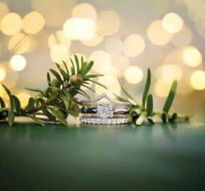 Ein silberner Ring mit einem funkelnden Diamanten in der Mitte ruht auf einer grünen Oberfläche, akzentuiert durch einen kleinen Zweig mit Blättern. Sanfte, kreisförmige Lichter verstärken das elegante und festliche Gefühl und fangen die Essenz der Hochzeitsplanung in dieser bezaubernden Szene ein.