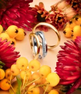 Drei ineinander verschlungene Ringe, darunter ein goldener und ein silberner Ring mit Diamanten, bilden ein atemberaubendes Herzstück inmitten leuchtend roter Chrysanthemen und Büschel kleiner gelb-oranger Beeren auf beigem Hintergrund – die perfekte Inspiration für alle, die sich gerade mit der Hochzeitsplanung beschäftigen.