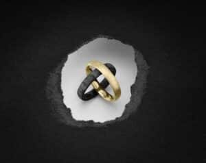 Zwei ineinander verschlungene Ringe, einer aus Gold und einer aus Dunkelgrau, liegen auf einem weißen, zerrissenen Papierhintergrund vor einer strukturierten schwarzen Oberfläche – perfekte Symbole für alle, die sich intensiv mit der Hochzeitsplanung beschäftigen.