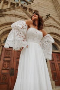 Eine Frau in einem schulterfreien weißen Spitzenkleid steht selbstbewusst vor einem Steingebäude mit Holztüren und verkörpert die Eleganz der Hochzeitsplanung. Sie blickt nach oben, ihre Arme ruhen auf ihren Hüften und heben die aufwendigen Spitzenärmel ihres Hochzeitsensembles hervor.