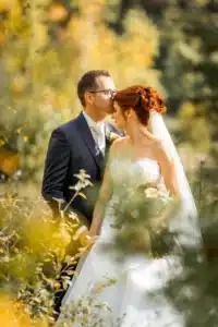 In einem sonnenbeschienenen Wald stehen Braut und Bräutigam an ihrem besonderen Hochzeitstag umgeben von Grün. Der Bräutigam küsst die Stirn der Braut, während sie ihren Blumenstrauß hält, beide lächeln. In einem weißen Kleid und Schleier gekleidet, ergänzt sie seinen dunklen Anzug wunderschön inmitten ihrer perfekten, liebevollen Umgebung.