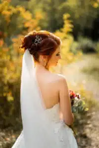 Eine Braut mit roten Haaren steht an ihrem Hochzeitstag in einem sonnenbeschienenen Garten. Sie trägt ein trägerloses weißes Kleid und hält einen Blumenstrauß in der Hand. Ihr Haar ist mit einer eleganten Spange gestylt und ein langer Schleier fällt ihr über den Rücken. Der Hintergrund ist mit Grünpflanzen verschwommen.