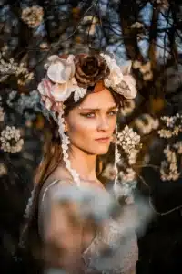 Eine Frau mit Blumenkopfschmuck steht zwischen blühenden Zweigen und weckt ein Gefühl von Hochzeitszauber. Sonnenlicht erhellt sanft ihr Gesicht und hebt ihr Make-up mit Glitzerdetails um die Augen hervor. Der Hintergrund zeigt verschwommene weiße Blüten und dunkle Zweige, perfekt für jede Hochzeitsplanungsinspiration.