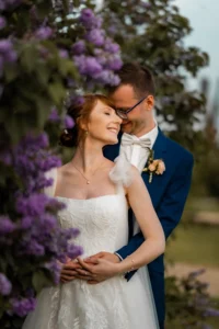 Braut und Bräutigam stehen eng beieinander und lächeln freudig inmitten üppiger lila Blumen. In ihrem weißen Hochzeitskleid umarmt sie ihren Bräutigam in einem blauen Anzug und einer Blumenboutonniere und fängt damit die Essenz einer perfekten Hochzeit in einer ruhigen Umgebung im Freien ein.