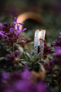 Nahaufnahme von zwei Diamantringen aus Roségold, perfekt für eine Hochzeit, eingebettet zwischen lila Blumen und Grünpflanzen. Die Ringe sind mit funkelnden Steinen geschmückt und auf einem von ihnen ruht eine kleine Blume. Ein ideales Symbol für Ihre Hochzeitsreise.