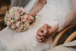 Eine Person im Hochzeitskleid hält einen Strauß rosa Rosen, sitzt mit gefalteten Händen da und zeigt zarte Spitzendetails auf dem Kleid. Die Blumen enthalten Schleierkraut und betonen einen ruhigen und intimen Hochzeitsmoment.