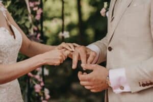 Braut und Bräutigam tauschen während ihrer Hochzeit im Freien die Ringe aus. Das weiße Spitzenkleid der Braut und der beige Anzug des Bräutigams ergänzen das üppige Grün und die rosa Blumen und schaffen eine bezaubernde Kulisse, die ihre Verbindung perfekt symbolisiert.