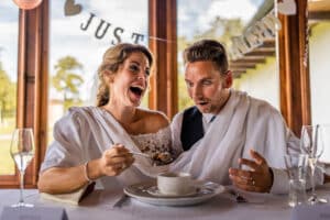 Ein fröhliches Brautpaar lacht an einem Empfangstisch, der mit einem „Just Married“-Banner geschmückt ist. Die Braut bietet dem Bräutigam spielerisch einen Löffel Essen an und verkörpert damit den Charme ihrer Hochzeit. Mit weißem Stoff drapiert, mit Gläsern und einem kleinen Tischgesteck ist es ein perfekter Moment inmitten ihrer Hochzeitsplanung.