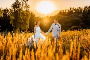 Braut und Bräutigam gehen bei Sonnenuntergang händchenhaltend durch ein Feld mit hohem Gras und verkörpern damit die Essenz ihrer Traumhochzeit. Die Sonne zaubert einen warmen, goldenen Schein um sie herum. Die Braut trägt ein fließendes weißes Kleid und der Bräutigam einen hellen Anzug mit Fliege. Im Hintergrund sind Bäume zu sehen.
