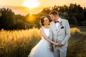 Ein Paar in Hochzeitskleidung steht bei Sonnenuntergang auf einem Feld und verkörpert die Essenz ihrer Hochzeit. Die Braut trägt ein weißes Kleid und hält die Hand des Bräutigams, der einen hellgrauen Anzug anzieht. Die Sonne geht hinter ihnen unter und wirft einen warmen Schein auf die Landschaft aus hohem Gras und Bäumen.