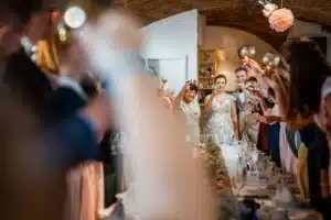 Braut und Bräutigam stehen in der Mitte eines langen, dekorierten Tisches und erheben ihre Gläser zum Hochzeitstoast. Gäste auf beiden Seiten spiegeln die Geste mit freudigen Ausdrücken in diesem warm beleuchteten, eleganten Veranstaltungsort mit gewölbten Decken wider.