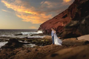 Ein Paar steht bei Sonnenuntergang an einem felsigen Strand und ist von der Romantik ihrer Hochzeit umgeben. Die Frau trägt ein fließendes weißes Kleid und der Mann einen Anzug. Dramatische Wolken bedecken den Himmel, während das Meer sanft gegen die Felsen schlägt und so eine perfekte Kulisse für ihre Liebesgeschichte bietet.