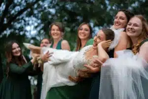 Eine fröhliche Braut in einem weißen Kleid und Schleier wird spielerisch von sechs Brautjungfern in dunkelgrünen Kleidern hochgehoben. Sie sind draußen, umgeben von Grün, feiern ihre Hochzeit mit einem Lächeln und blicken in die Kamera.
