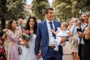 Braut und Bräutigam gehen Hand in Hand im Freien spazieren, umgeben von lächelnden Gästen. In dieser fröhlichen Hochzeitsszene wiegt der Bräutigam ein Baby, während die Braut einen Strauß rosa und weißer Blumen hält. Die Feier geht weiter, während Seifenblasen herumschweben, während im Hintergrund Bäume den Weg säumen.