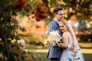 Braut und Bräutigam umarmen sich in einer Gartenkulisse, eingefangen in der Magie ihrer Hochzeit. Die Braut hält einen Strauß weißer Blumen und legt ihren Kopf auf die Schulter des Bräutigams. Sonnenlicht fällt durch die Bäume und wirft einen warmen, romantischen Schein auf diese bezaubernde Szene.
