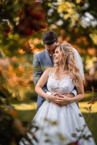 Inmitten einer malerischen Gartenkulisse umarmt sich ein Paar zärtlich; die Frau im Spitzenbrautkleid und der Mann im grauen Anzug verkörpern die Essenz des romantischen Heiratens. Sonnenlicht fällt durch das üppige Grün und verleiht ihrem perfekten Hochzeitsplanungsmoment Wärme.