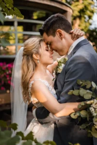 Braut und Bräutigam stehen dicht beieinander, die Augen geschlossen, die Stirnen berühren sich. Die Braut in weißem Kleid und Schleier umarmt den Bräutigam im grauen Anzug. Inmitten üppigen Grüns und sanften Lichts spiegelt ihr Moment die Schönheit der Hochzeitsplanung in ihrer reinsten Form wider.
