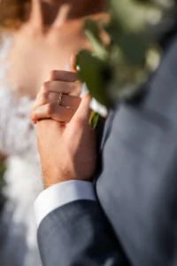 Nahaufnahme eines Paares, das sich an den Händen hält, mit Fokus auf die ineinander verschlungenen Finger. Die Braut in Spitzenkleid und Ehering und der Bräutigam im grauen Anzug teilen einen intimen Moment voller Wärme – ein perfekter Schnappschuss, der ihre sorgfältige Hochzeitsplanung widerspiegelt.