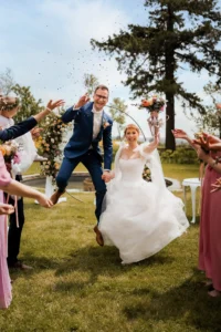 Ein fröhliches Paar feiert seine Hochzeit im Freien. Die Braut im weißen Kleid und der Bräutigam im blauen Anzug springen herum und halten Händchen. Die Gäste um sie herum werfen Konfetti und halten Blumen in den Händen, was zur festlichen Atmosphäre beiträgt.