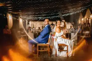 Braut und Bräutigam spielen bei ihrem Hochzeitsempfang das Schuhspiel. Sie sitzen Rücken an Rücken auf Holzstühlen, lachen und halten Schuhe hoch. Der Veranstaltungsort, wunderschön mit Lichterketten dekoriert, strahlt eine fröhliche Atmosphäre aus, während die Gäste diesen wunderbaren Moment im Hintergrund genießen.