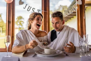 Braut und Bräutigam, in elegante weiße Tücher gekleidet, sitzen an einem Tisch. Die Braut füttert den Bräutigam mit einem Löffel Essen, beide zeigen lebhafte, überraschte Ausdrücke. Im Hintergrund feiert ein „Just Married“-Banner ihre Hochzeit und fängt diesen freudigen Moment ihrer Heiratsreise ein.