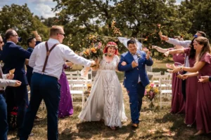Eine fröhliche Hochzeitsszene mit einer Braut in einem weißen Kleid und einem Bräutigam in einem blauen Anzug, die draußen den Gang entlanggehen. Die Gäste feiern, indem sie Konfetti werfen, mit Grünpflanzen und Blumenarrangements im Hintergrund, die die Essenz perfekter Hochzeitsplanung einfangen.