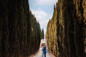 Ein Paar umarmt sich auf einem von hohen, dichten Bäumen gesäumten Weg und verkörpert die romantische Gelassenheit ihres Hochzeitstages. Der Bräutigam im blauen Anzug und die Braut im weißen Kleid stehen unter einem teilweise bewölkten Himmel und fangen die Essenz dieses wunderschön geplanten Moments ein.