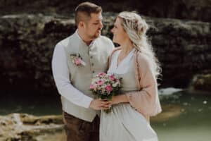 Ein Paar in Hochzeitskleidung steht an einem felsigen Flussufer. Der Mann in der Weste hält die Frau an der Taille, während sie in einem weißen Kleid und einem Schal einen Strauß rosa Blumen hält. In diesem perfekten Moment ihrer Hochzeitsreise blicken sie sich lächelnd in die Augen.
