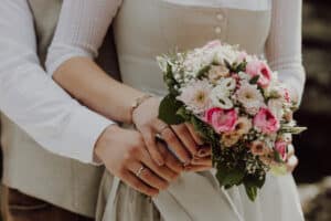 Eine Nahaufnahme eines Paares, das sich an den Händen hält. Die Frau im weißen Kleid hält einen Strauß rosa und weißer Blumen in der Hand. Die Hand des Mannes ruht sanft auf ihrer und fängt die Essenz ihrer Hochzeit ein. Die Kulisse erinnert auf wunderschöne Weise an eine Hochzeit oder einen formellen Anlass, bei dem Liebe und Verbundenheit gefeiert werden.